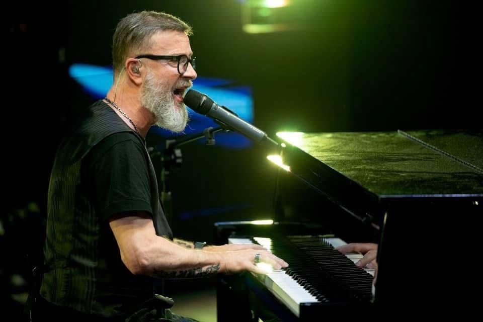 Un musicista canta in un microfono mentre suona il pianoforte su un palco buio.