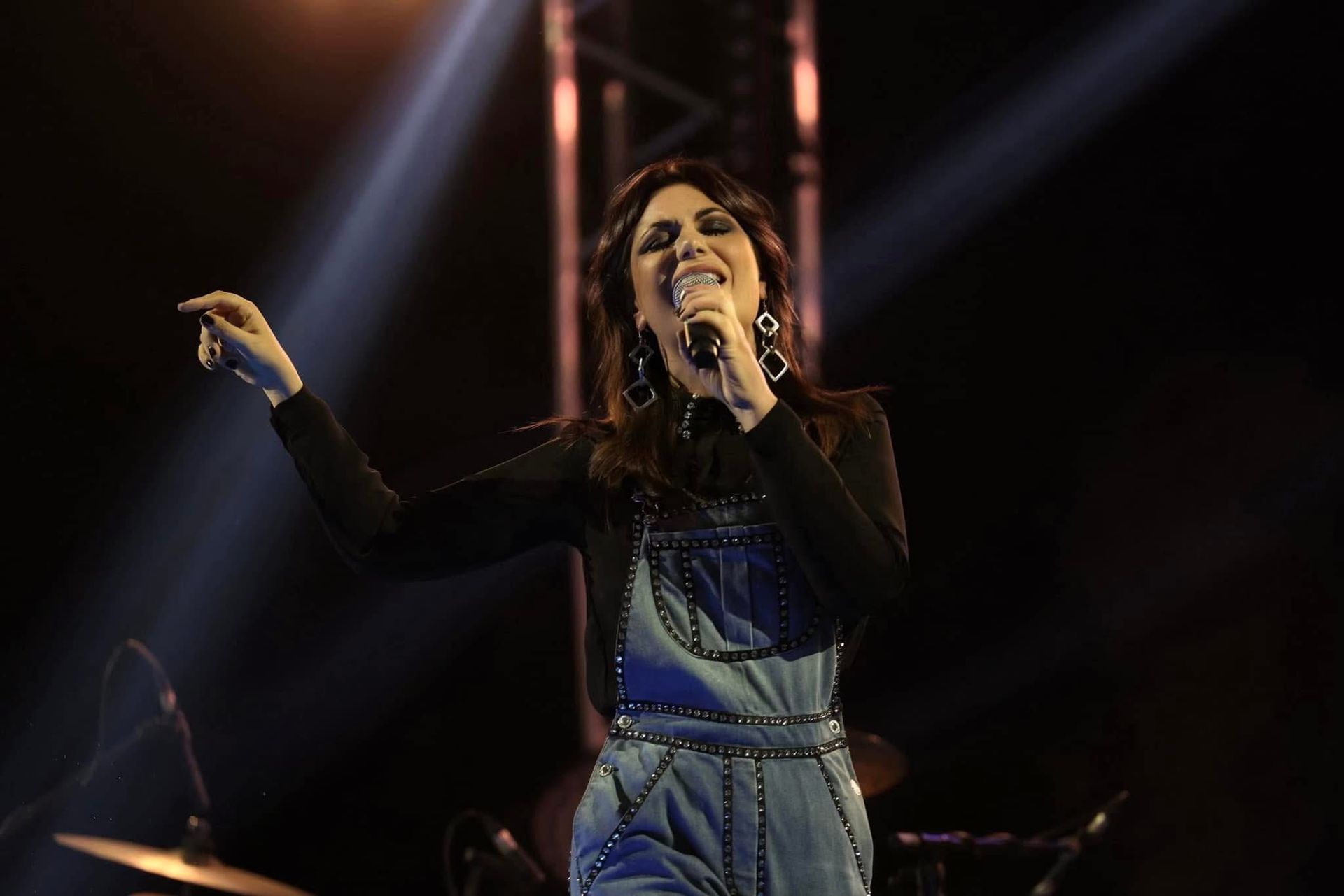 Cantante sul palco con un microfono in mano sotto i riflettori, che indossa una tuta di jeans e una maglietta nera.