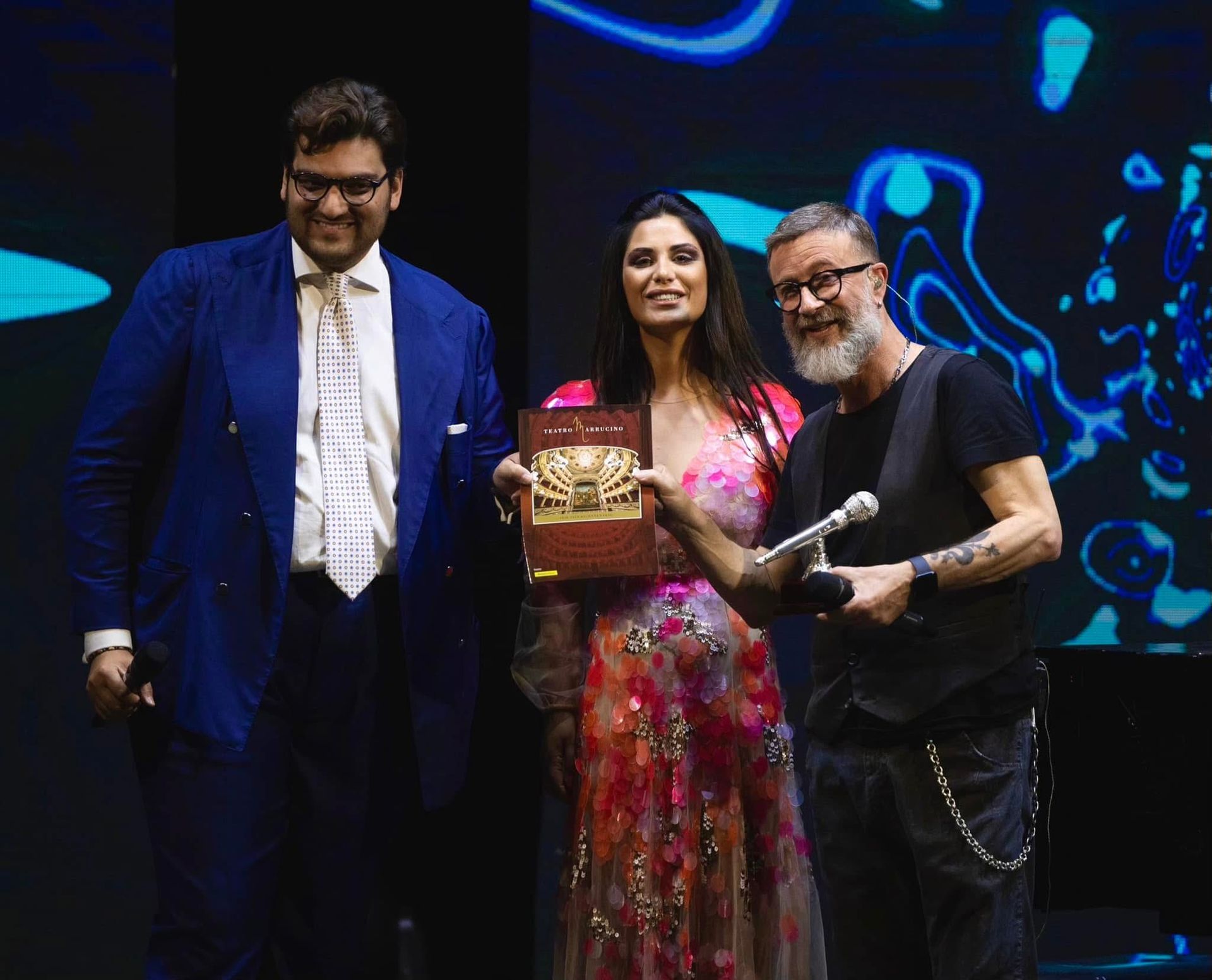 Tre persone sul palco sorridenti, che tengono in mano un trofeo e un microfono, con uno sfondo colorato.