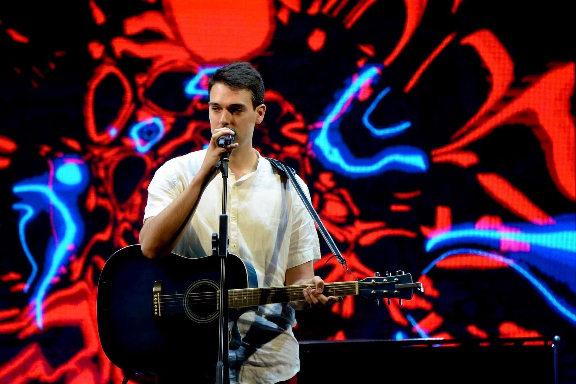 Cantante che si esibisce sul palco con la chitarra acustica davanti a uno sfondo astratto rosso e blu.