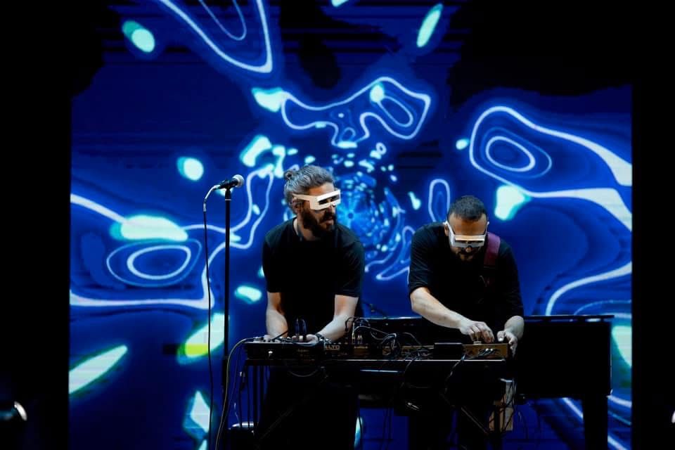 Due DJ si esibiscono a una consolle sotto luci al neon blu e uno sfondo astratto a forma di onda.