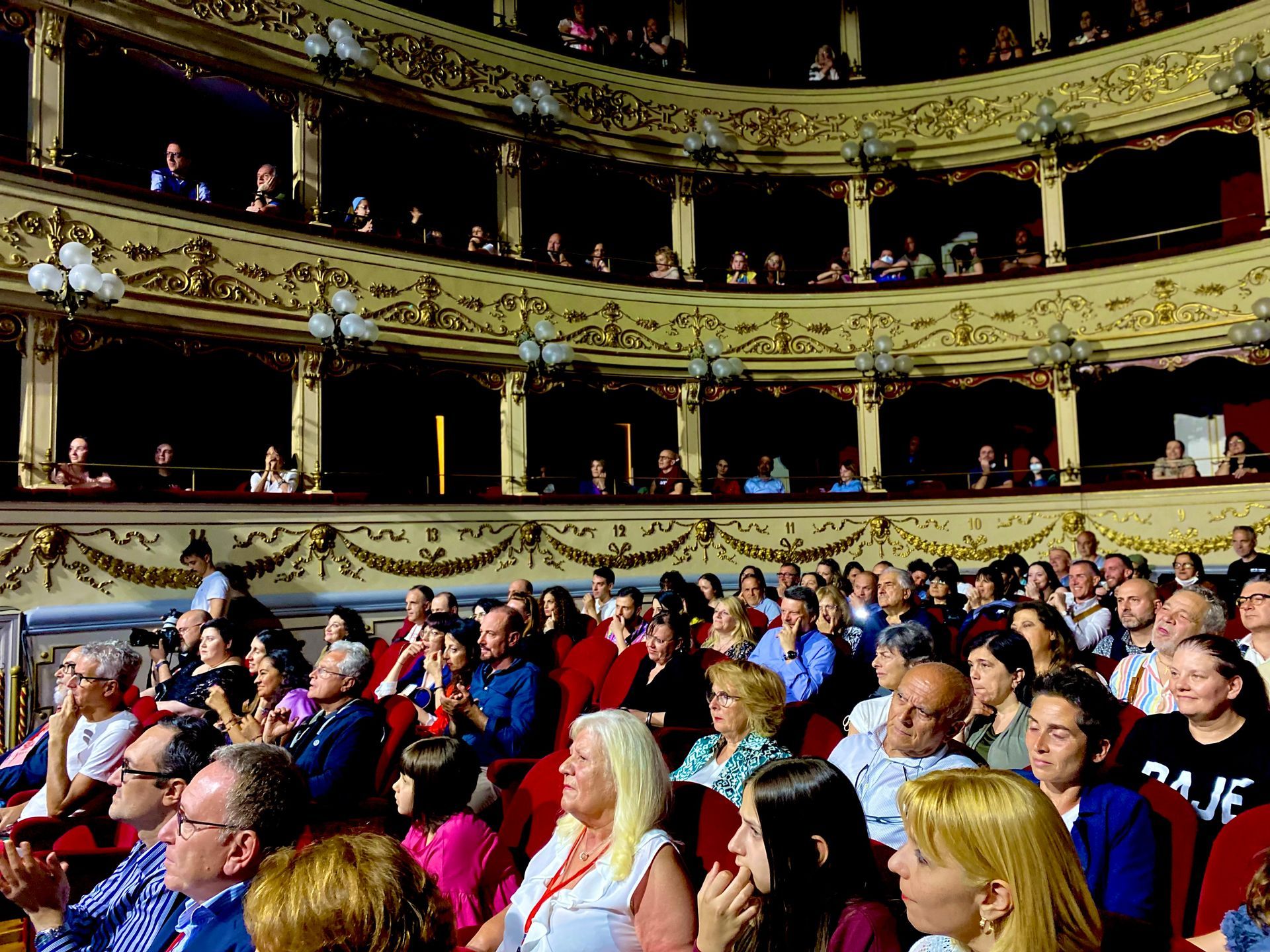 Pubblico seduto su una galleria teatrale riccamente decorata, intento ad assistere a uno spettacolo.