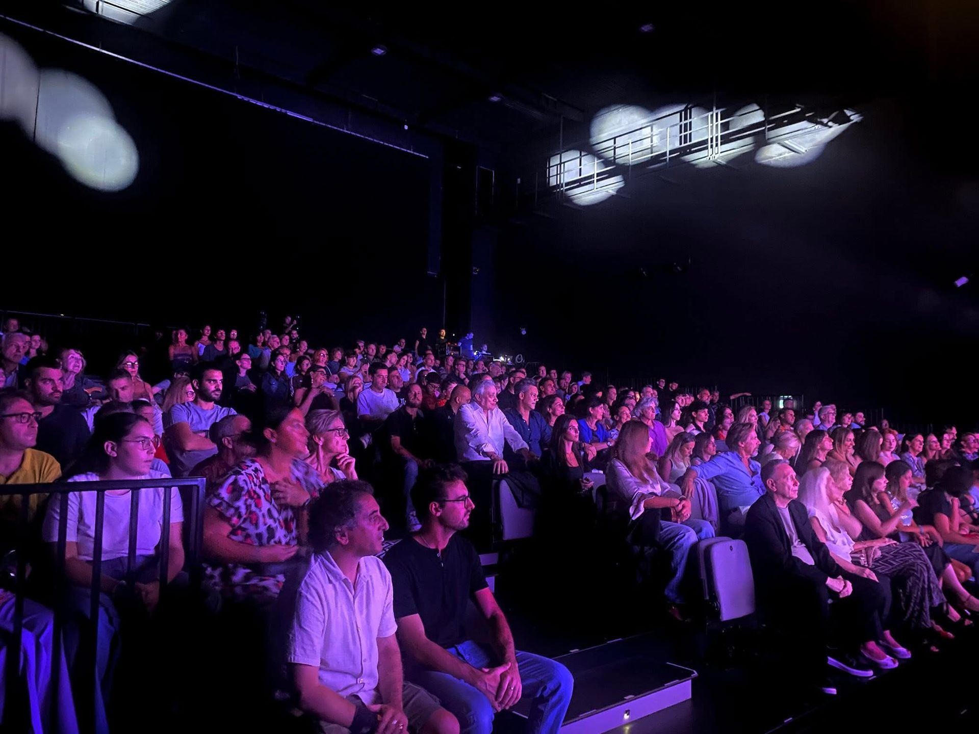 Il pubblico seduto in un teatro scarsamente illuminato osserva un palcoscenico illuminato da luci viola.