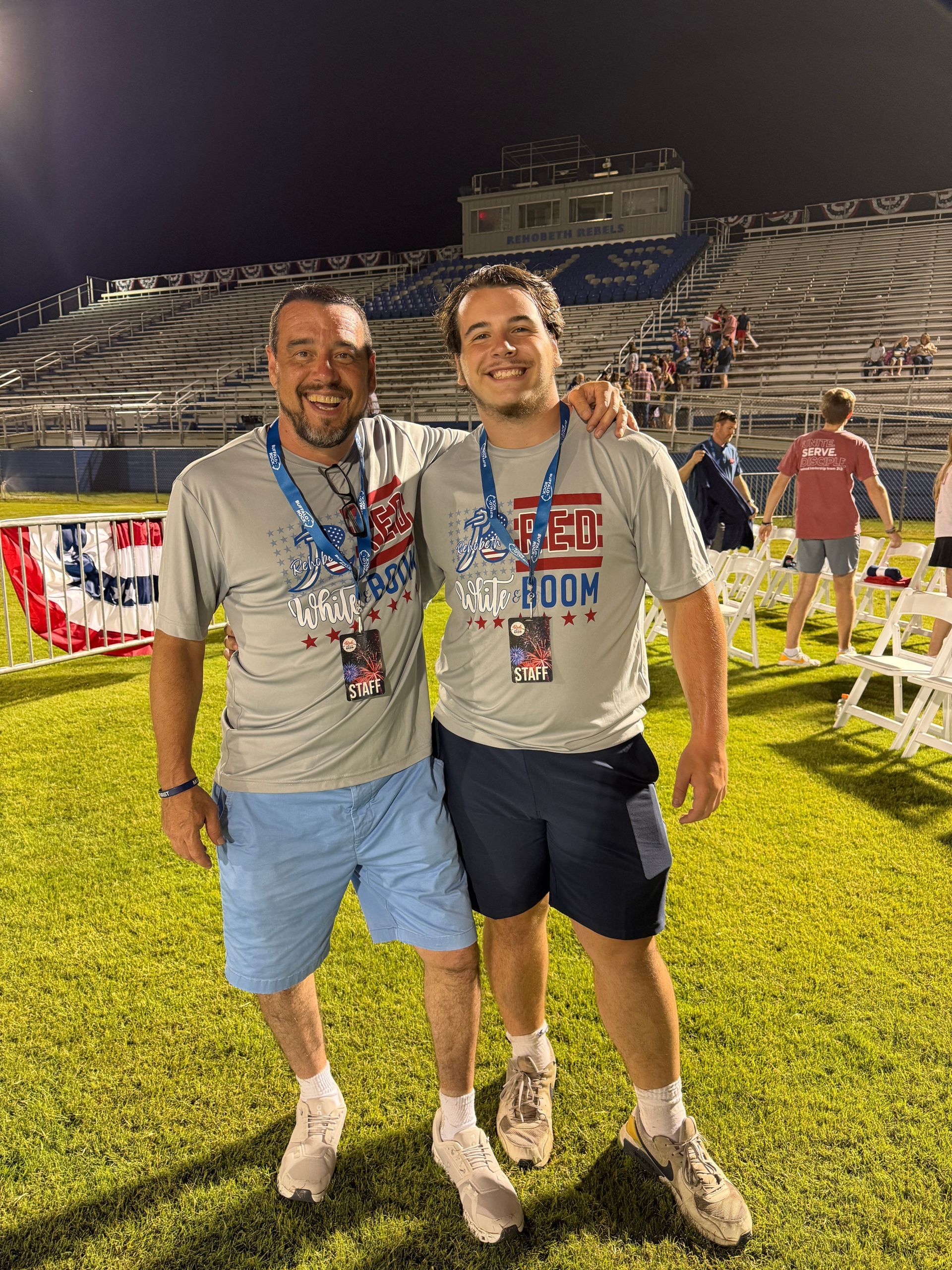 Two men with arms around each other, smiling, wearing matching shirts and lanyards, on a field with bleachers in background.