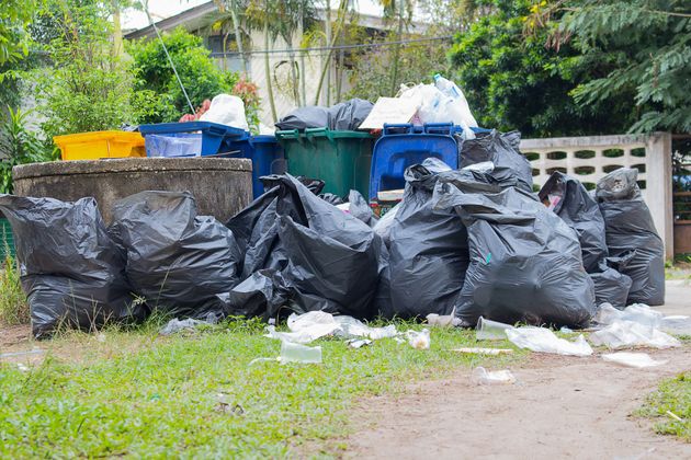 Black garbage bags piled along a curb beside blue bins and a yellow container in a grassy area