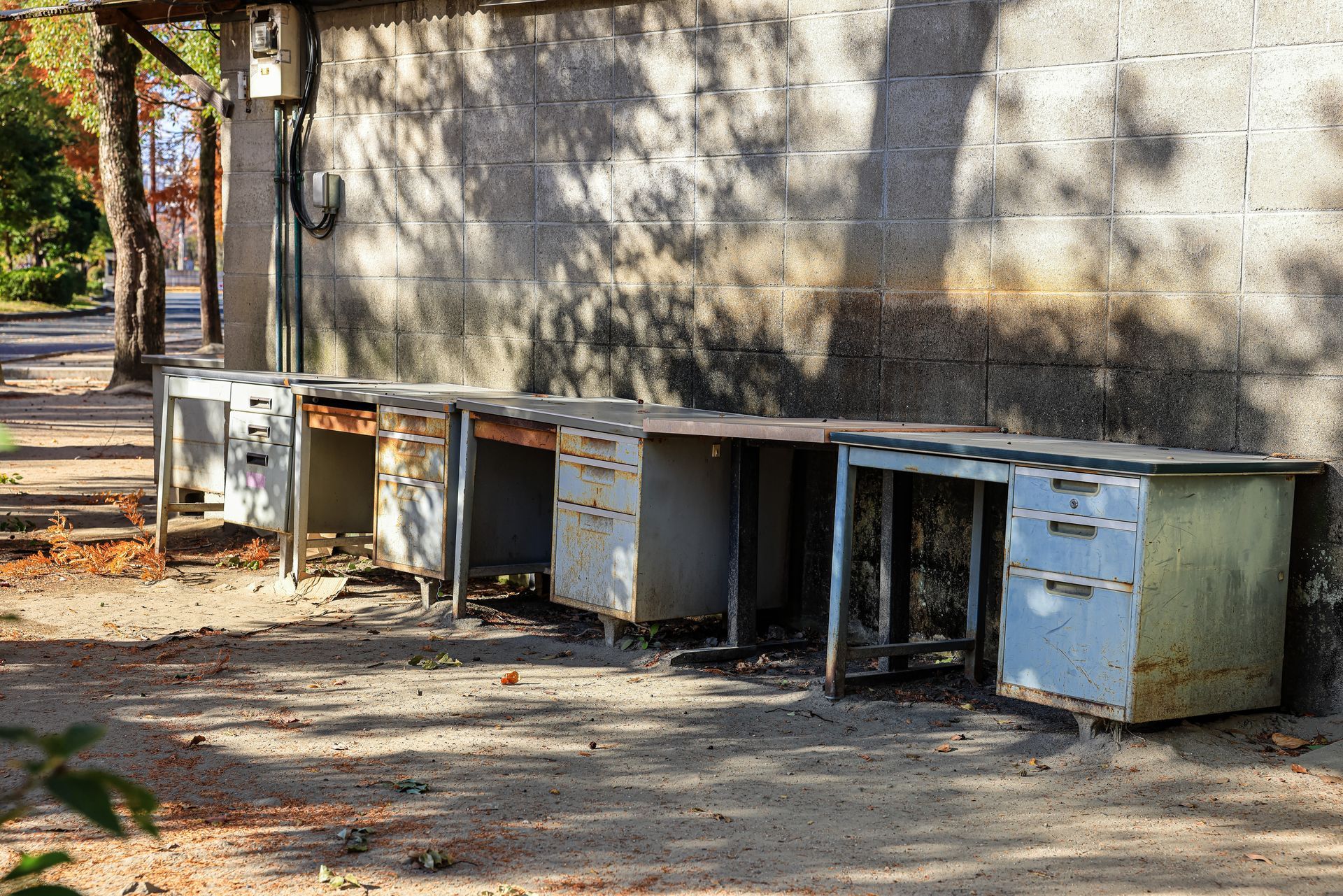 Old metal filing cabinets lined up outdoors beside a weathered wall in a yard with tree shadows.