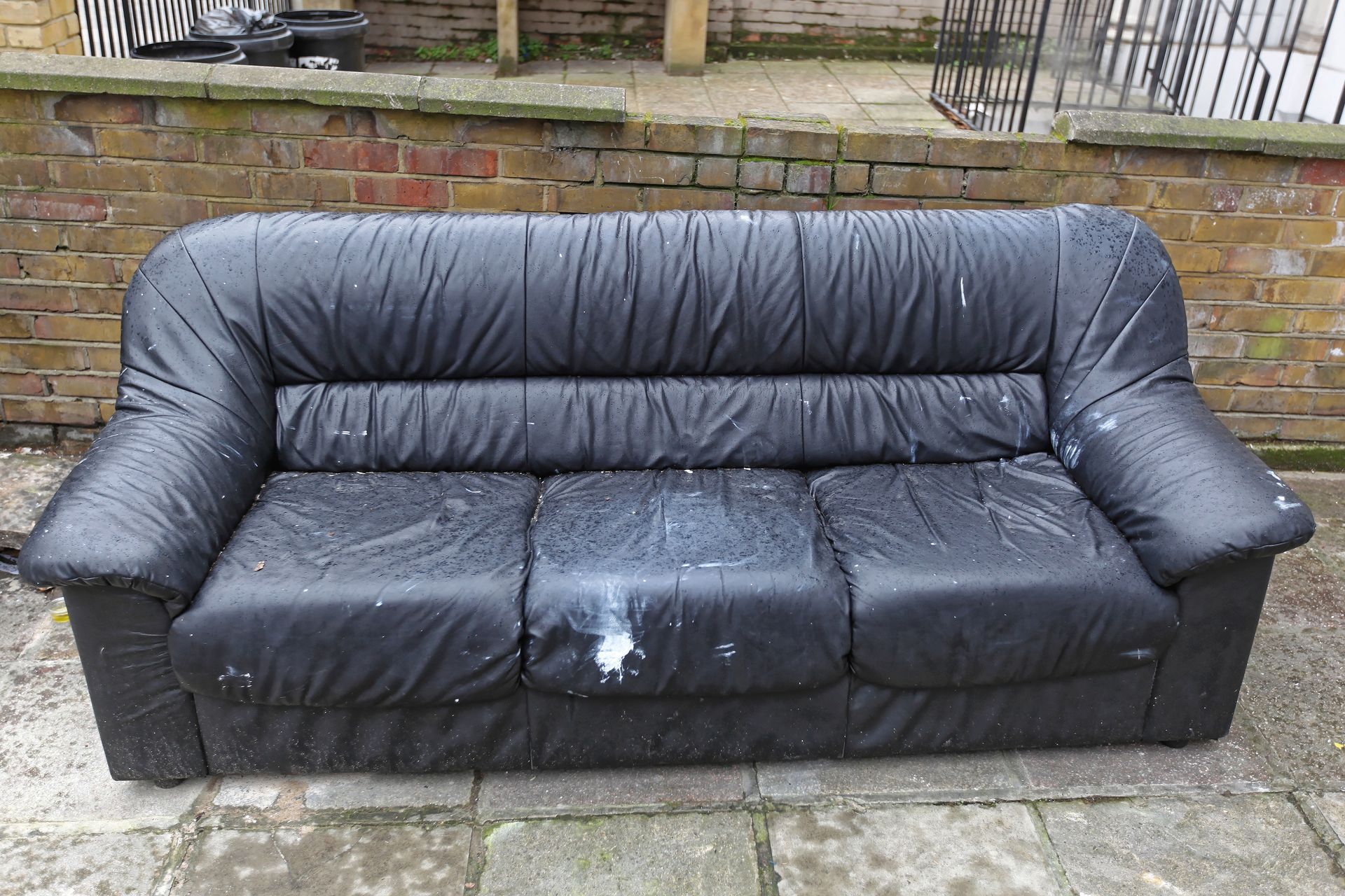 Black sofa with wet stains sitting on a stone patio against a brick wall