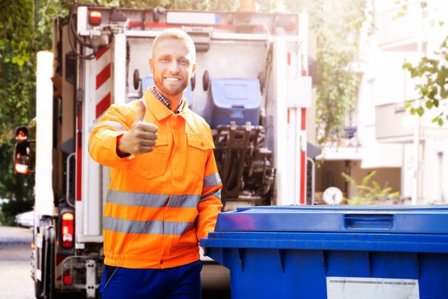 Smiling sanitation worker in orange reflective jacket giving thumbs up beside a garbage truck and blue bin