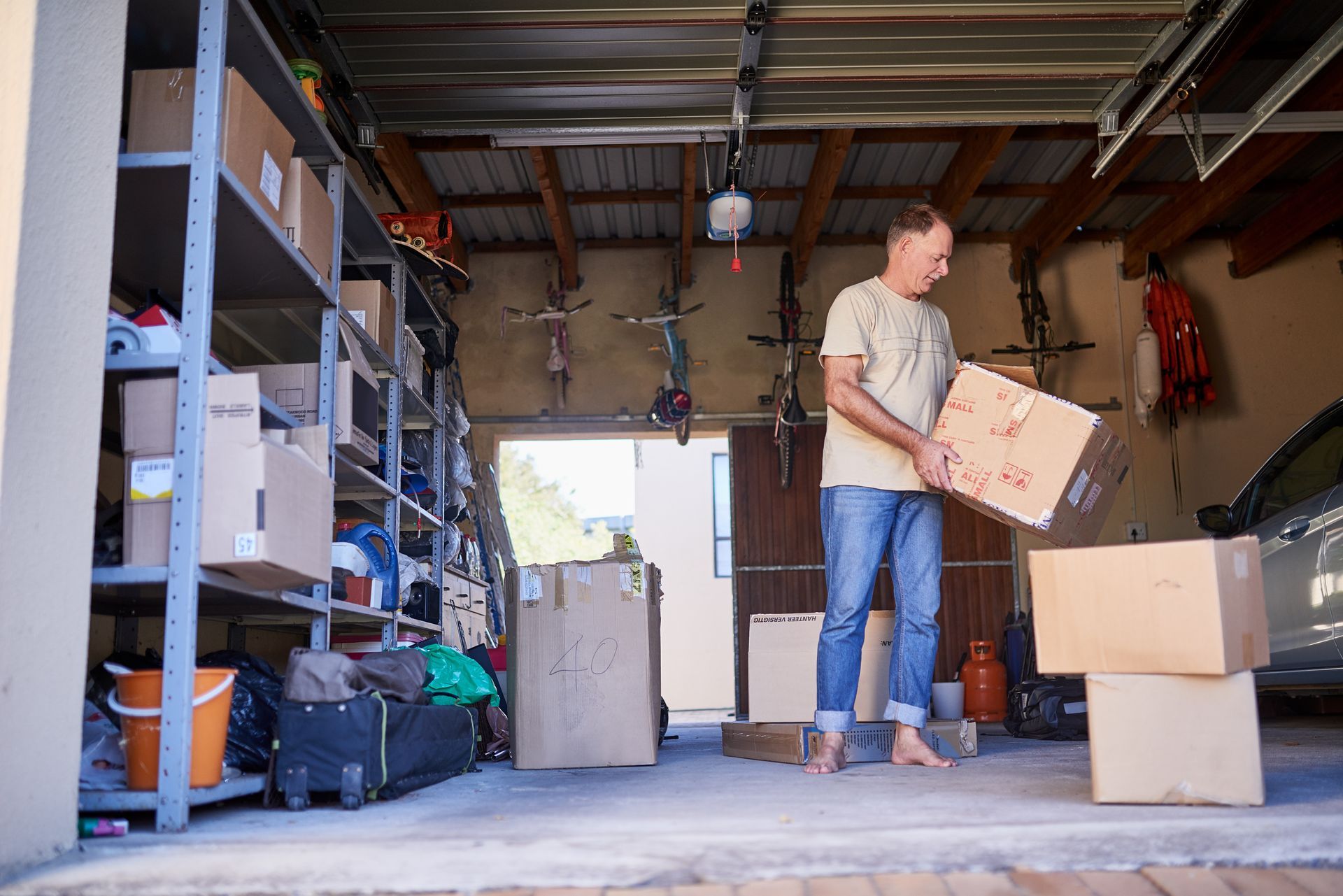 Person carrying a cardboard box in a garage filled with storage shelves and moving boxes.
