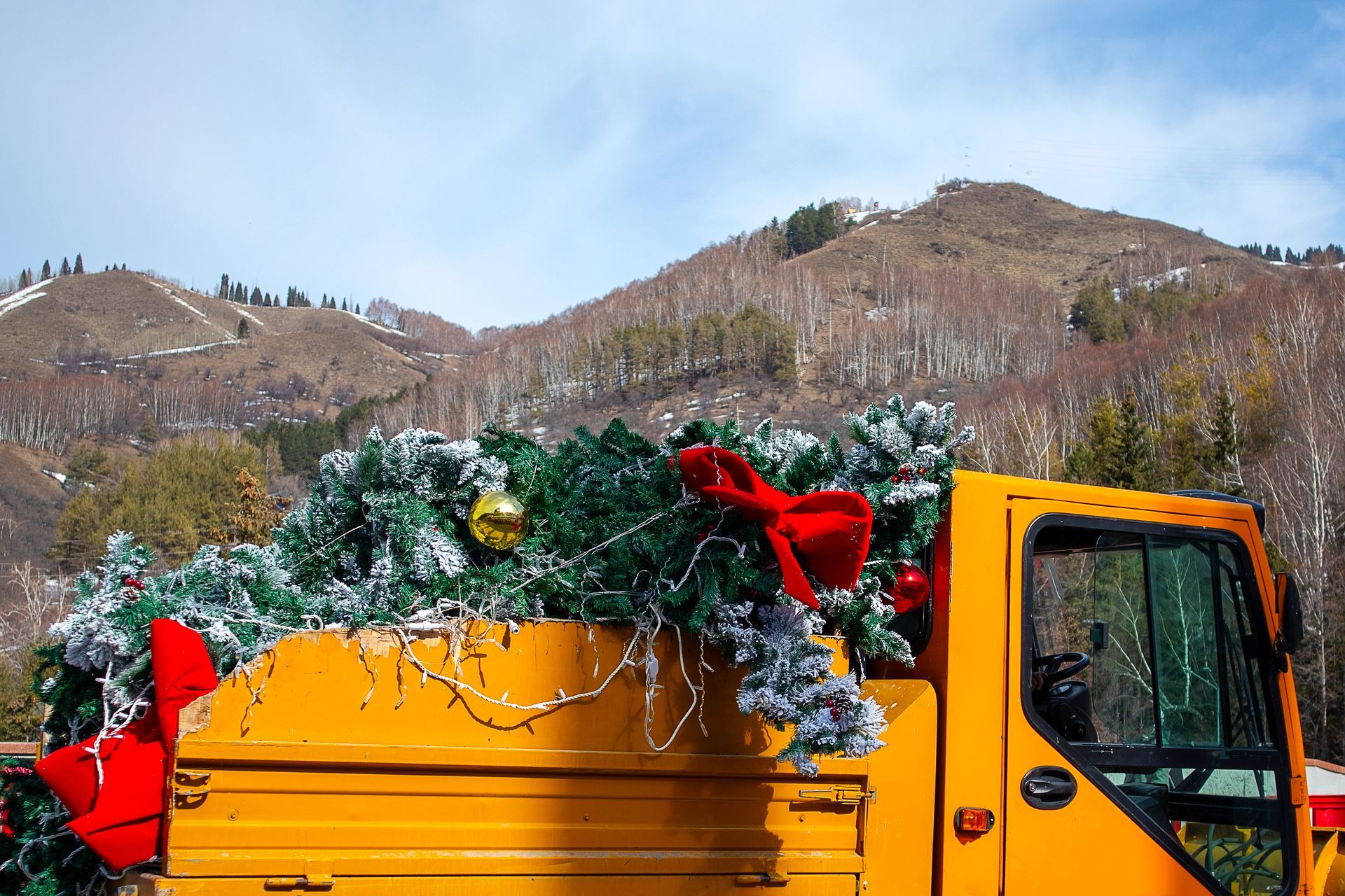 Yellow dump truck loaded with holiday greenery and a red bow in a snowy mountain landscape