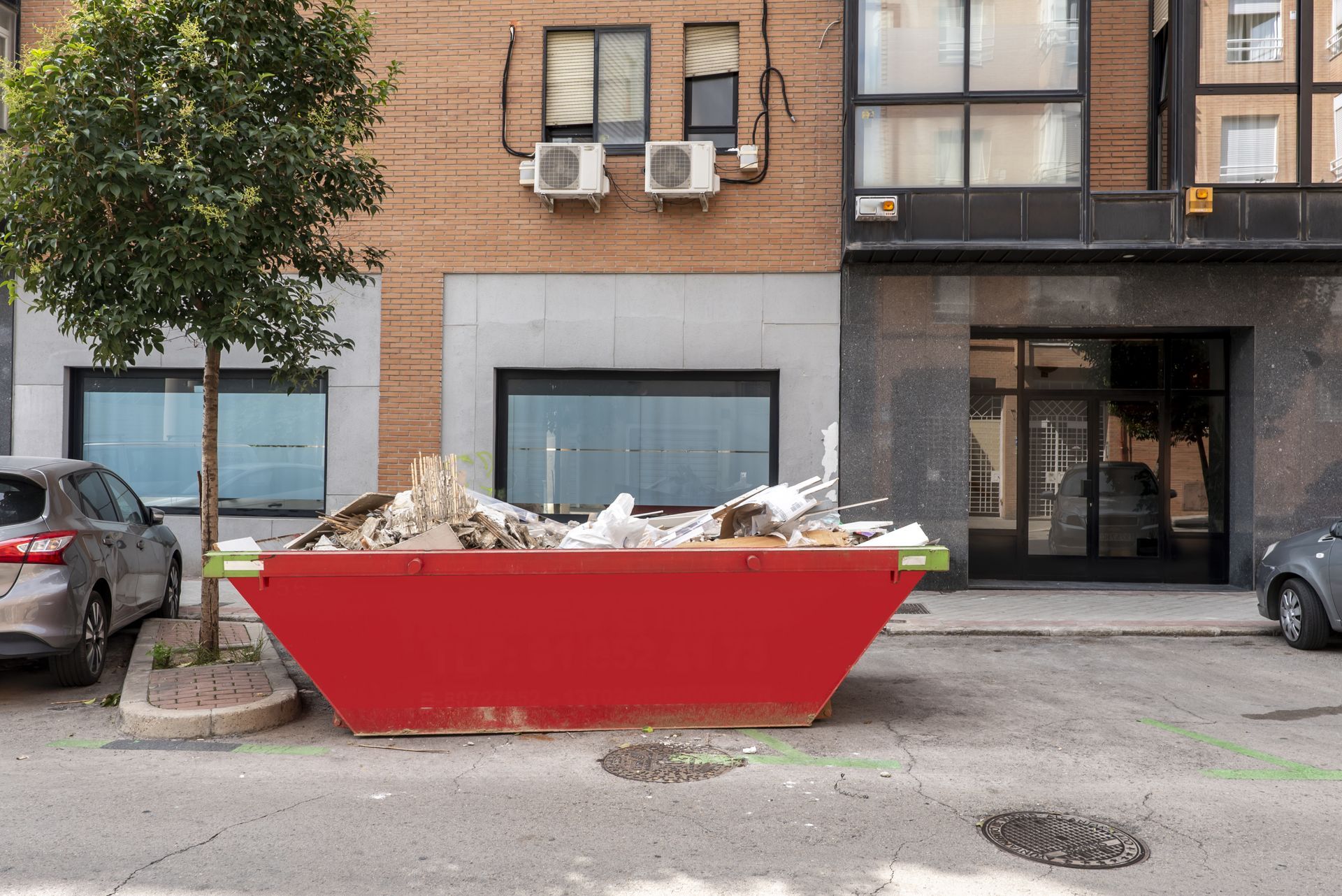 Red dumpster filled with debris on a city street in front of brick storefronts