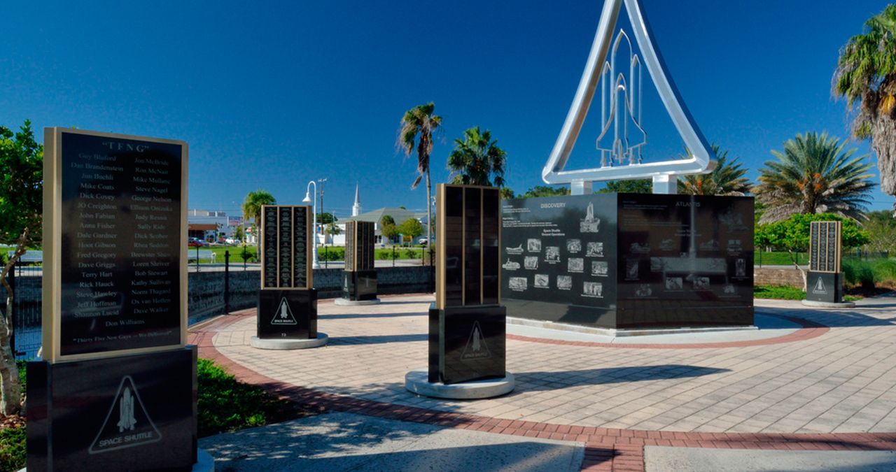 Outdoor memorial plaza with black stone monuments, palm trees, and a blue triangular sculpture under a clear sky