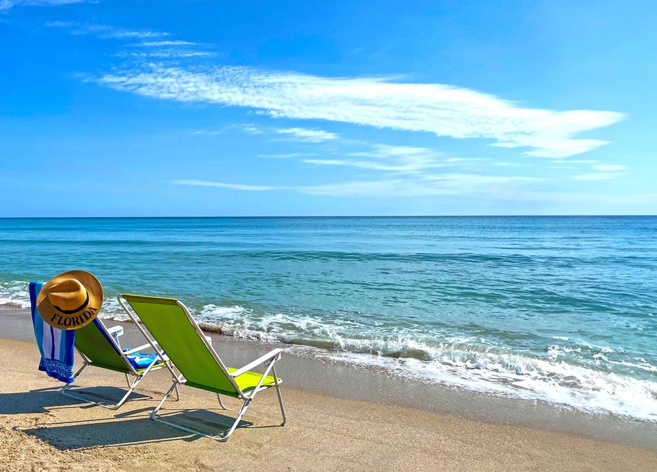 Two beach chairs and a sun hat on a sandy shore beside calm blue ocean under a bright sky