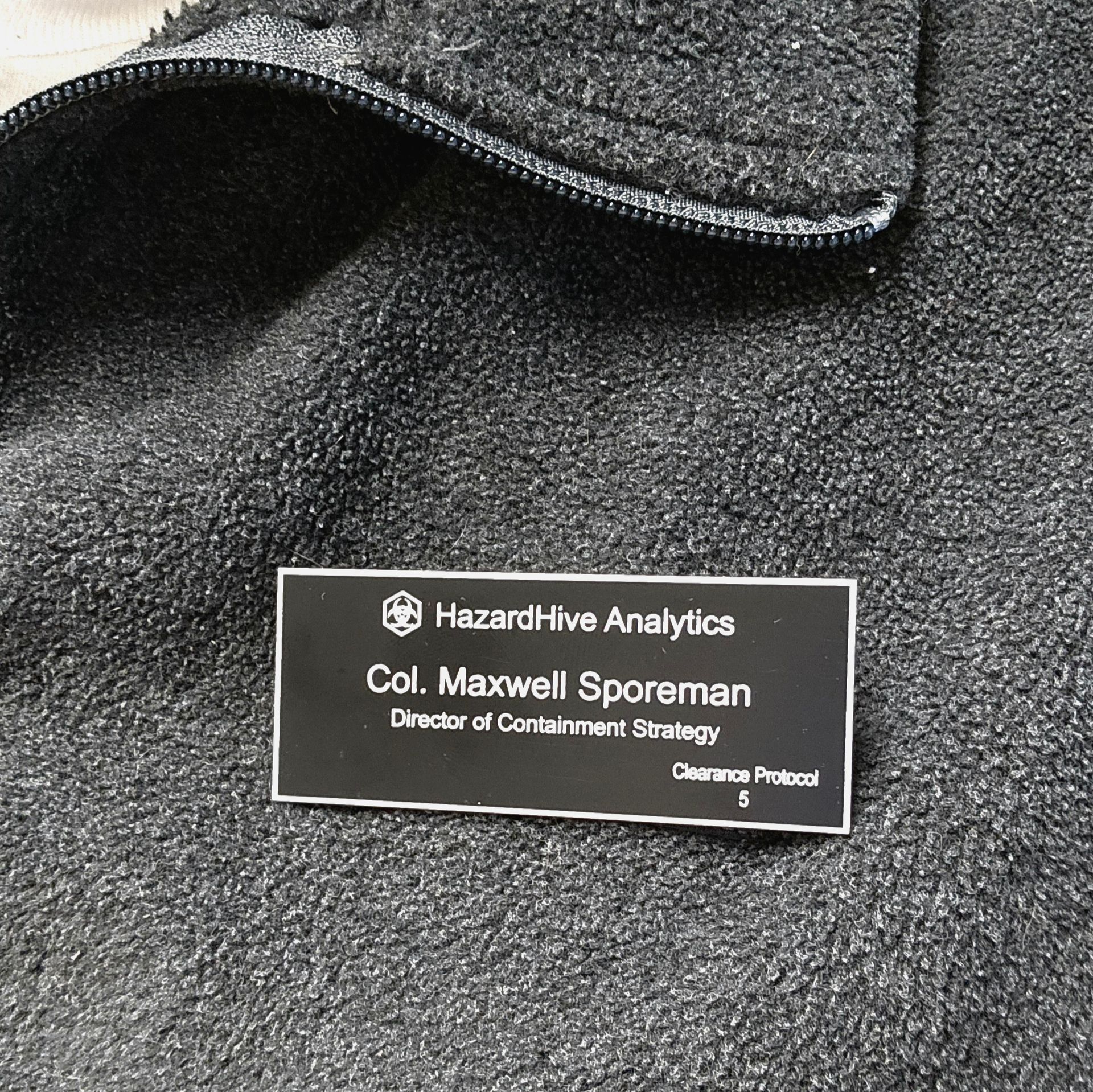 Black engraved name badge with white text attached to gray fleece jacket

