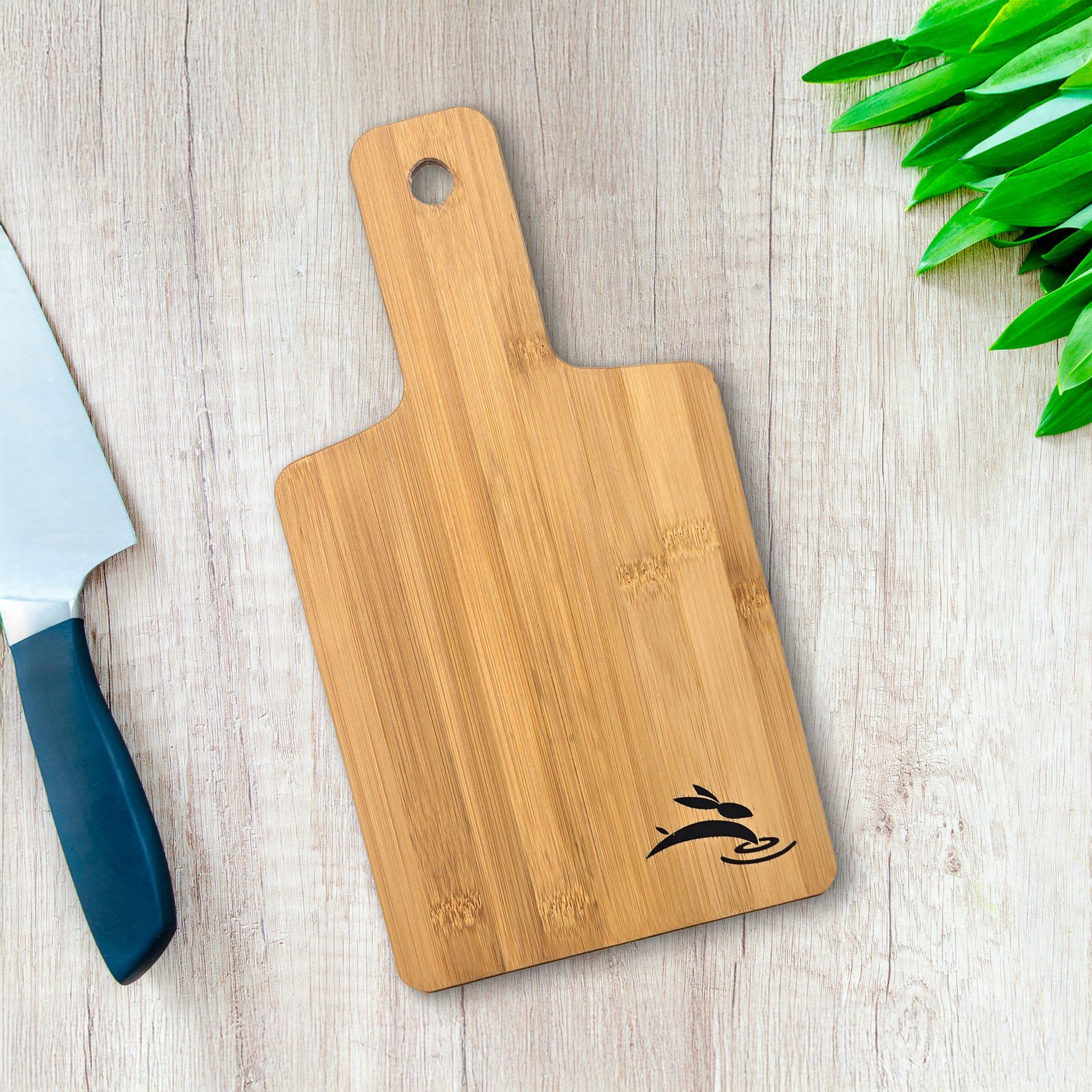 Bamboo cutting board mockup with engraved logo on wood surface beside kitchen knife