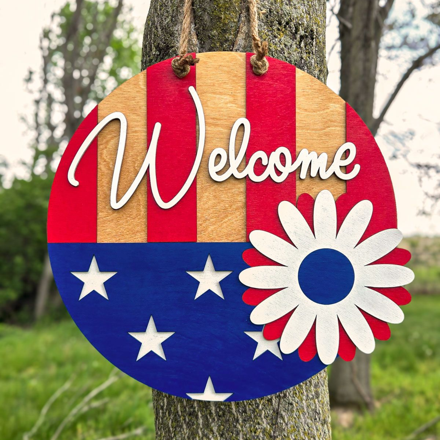 Layered patriotic welcome sign with stars, stripes, and flower hanging on tree
