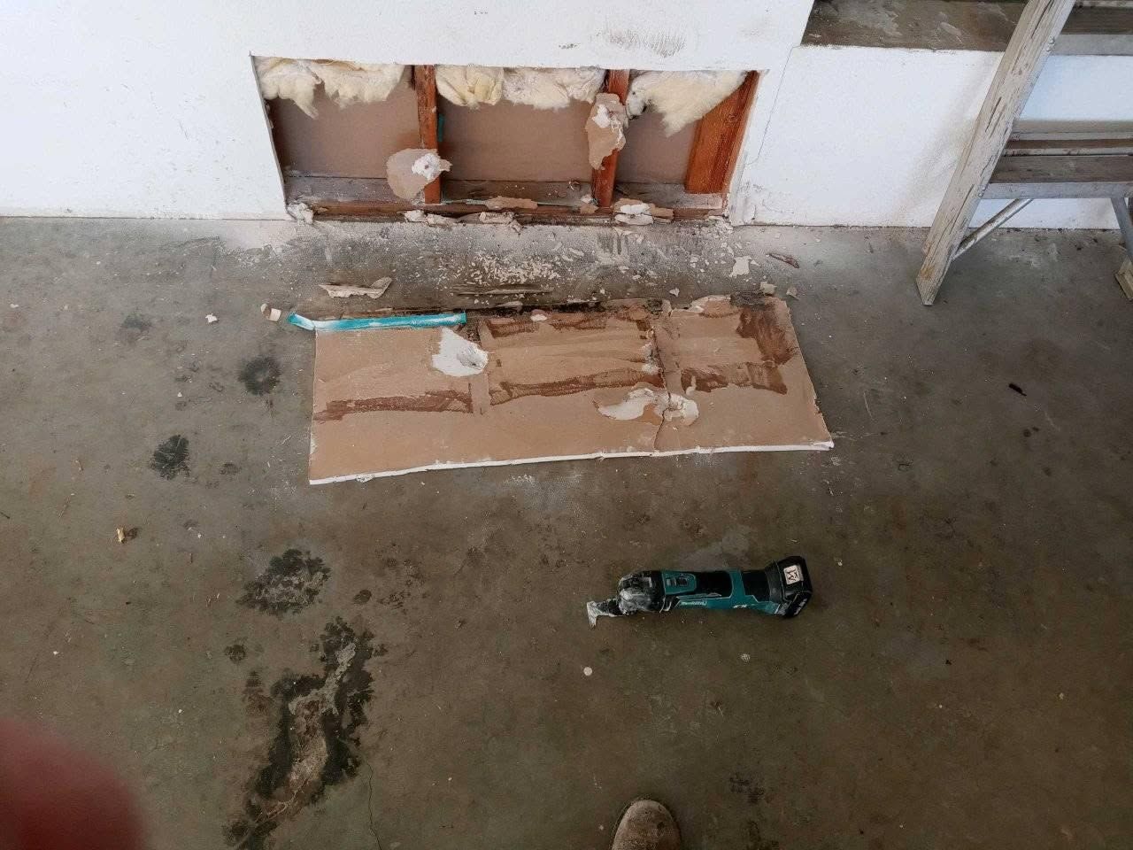 Damaged wall with exposed pipes and a loose cardboard panel on a concrete floor; hand tool nearby