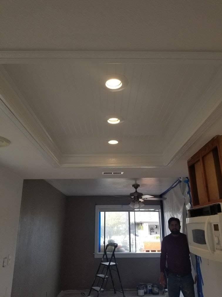 Living room under renovation with recessed ceiling lights, a window, ladder, fan, and a person standing by the wall