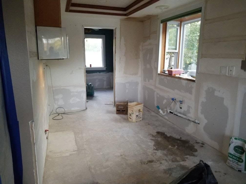 Renovation work in a narrow room with unfinished walls, dusty floor, and windows on the right.