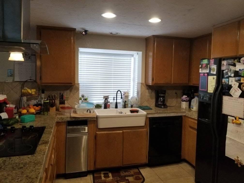 Kitchen with wooden cabinets, white farmhouse sink under a window, and a black refrigerator with papers on it.