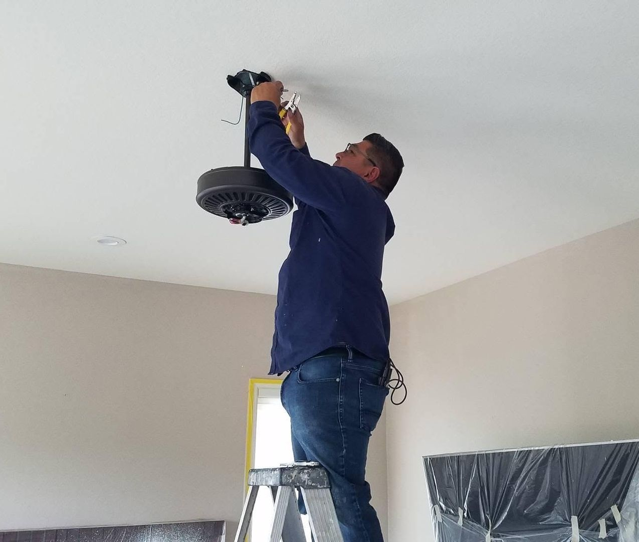 Person on a ladder installing a black ceiling fan in a beige room