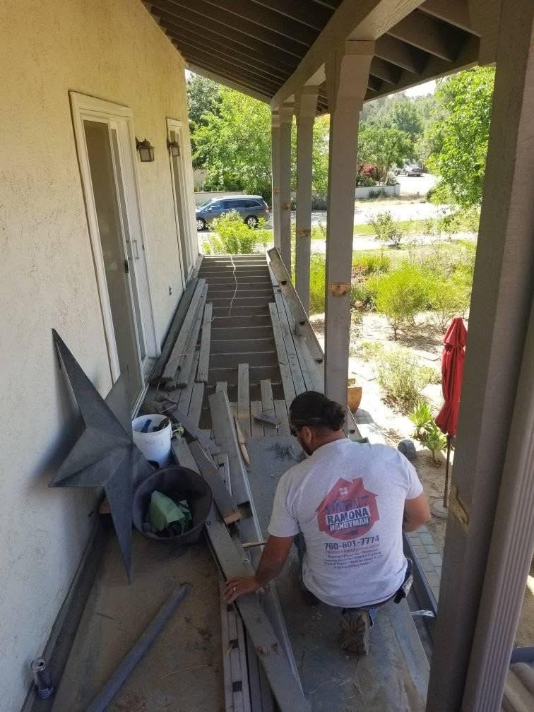 Person crouching on a narrow porch, working beside stacked boards and tools under a covered walkway.