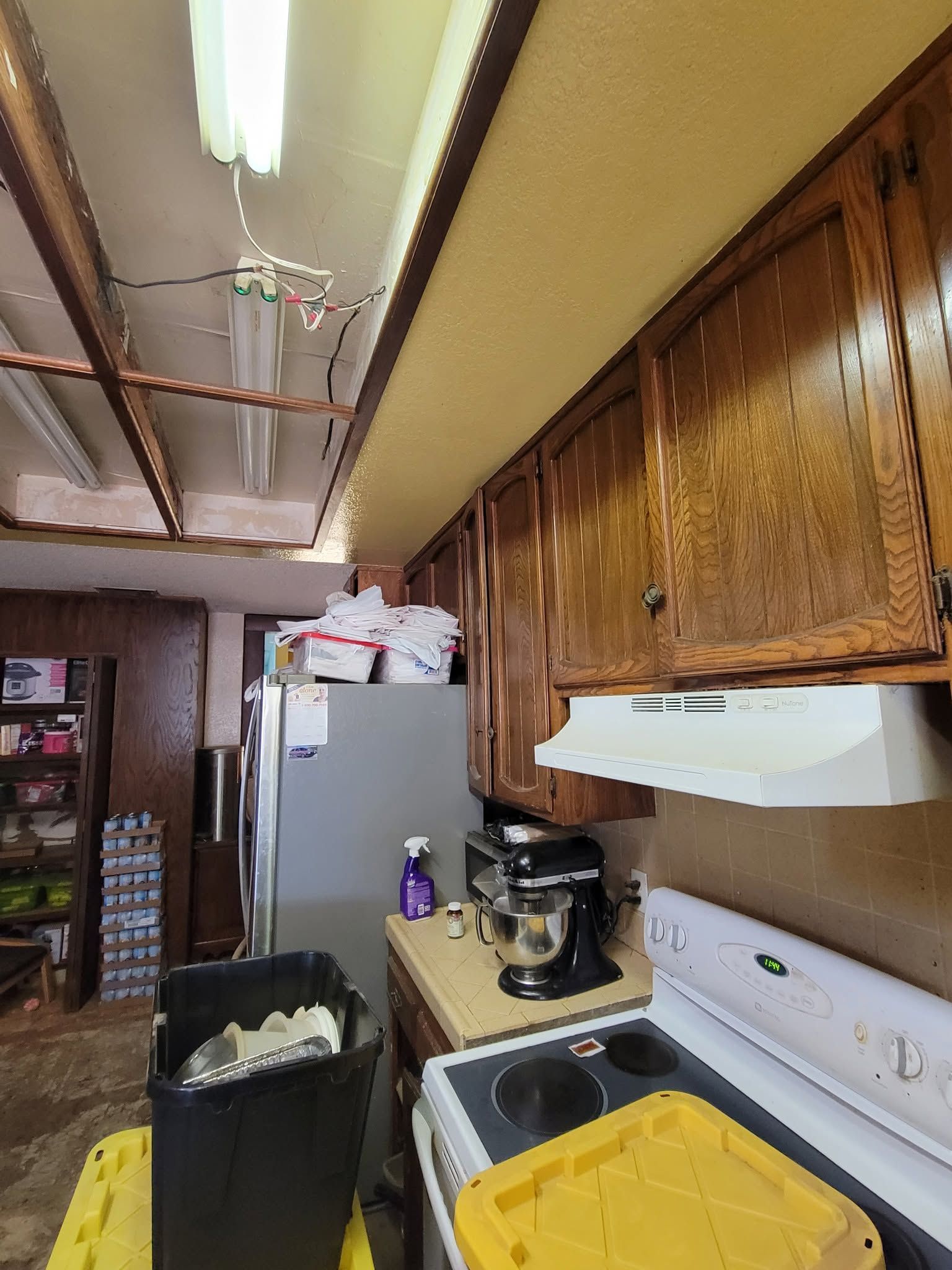 Narrow kitchen with wood cabinets, refrigerator, stove, and countertop appliances under fluorescent lighting