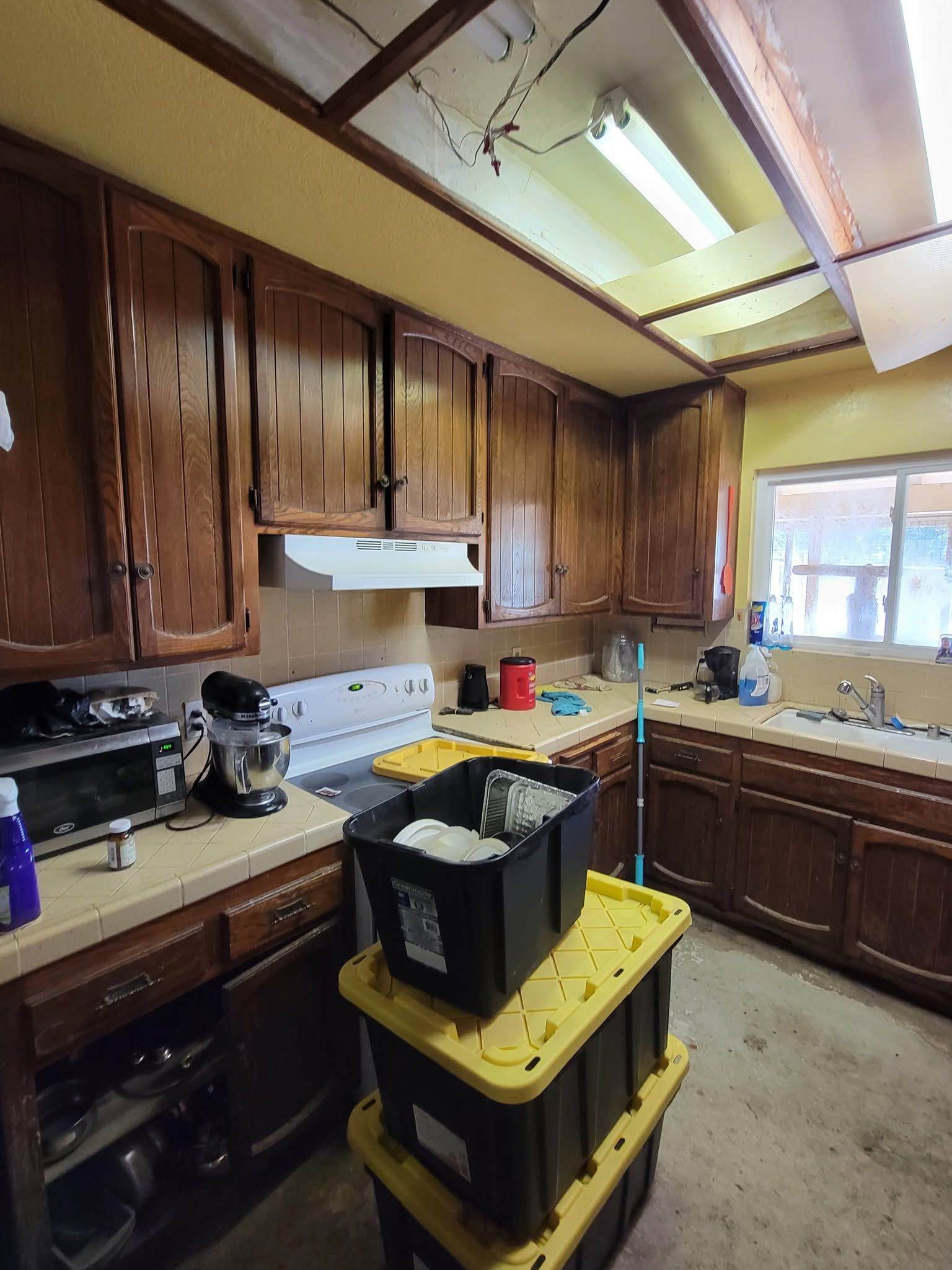 Cluttered kitchen with wooden cabinets, stove, sink, and stacked storage bins in the center.