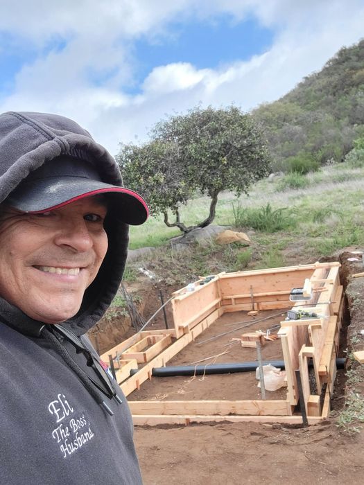 Smiling man in hooded jacket at a wooden construction site on a hillside under cloudy skies