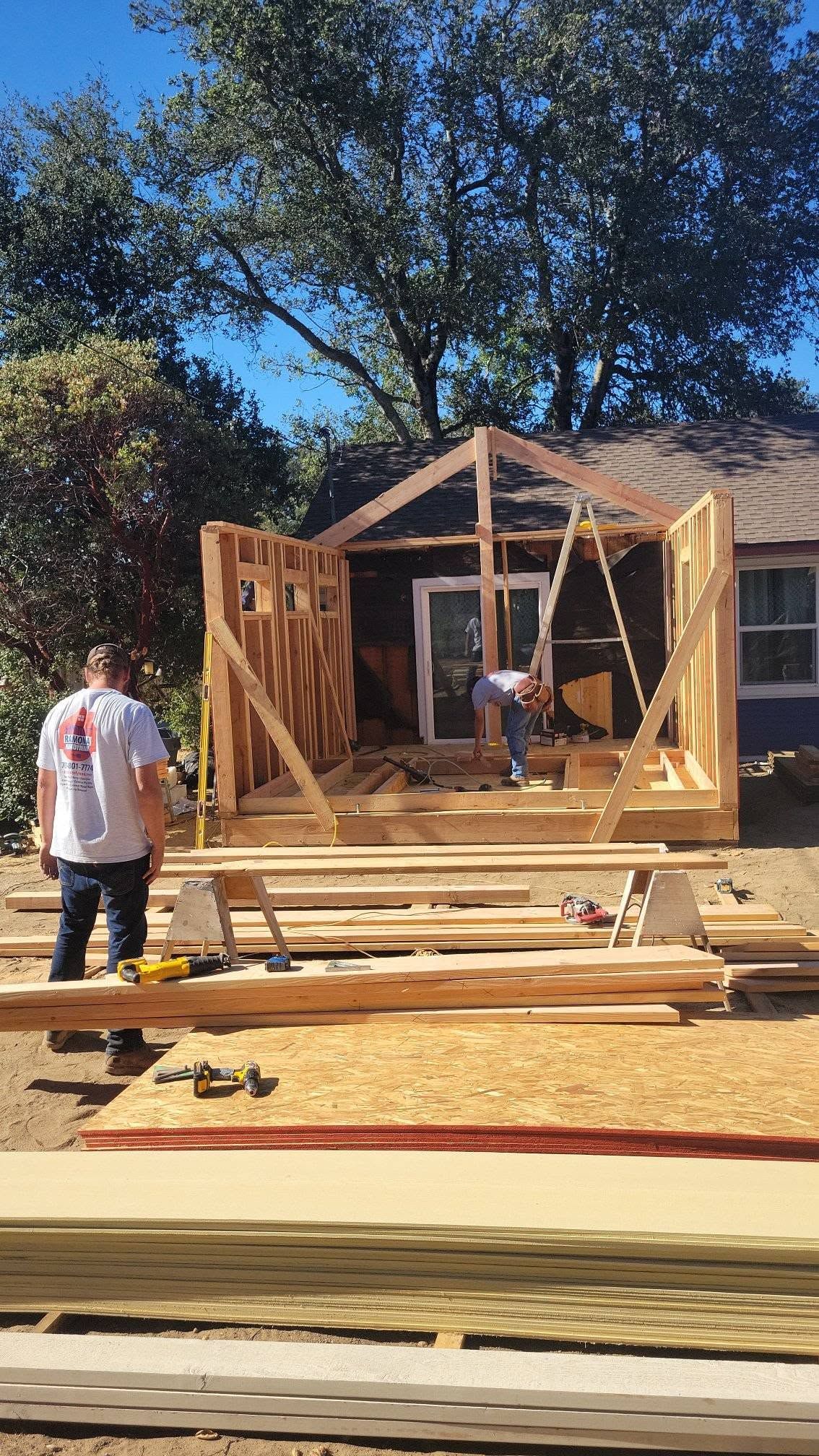 Workers framing a small house addition in a sunny backyard, with lumber stacked in front.