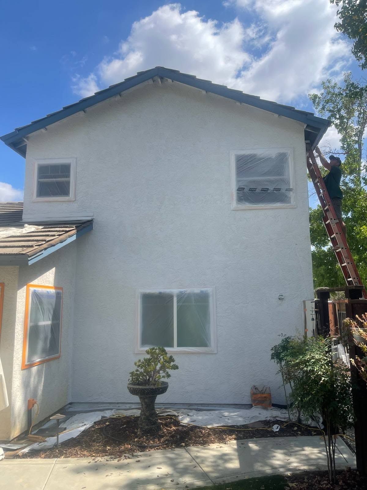 Two-story white stucco house with gray roof, blue sky, and a small front patio with plants.