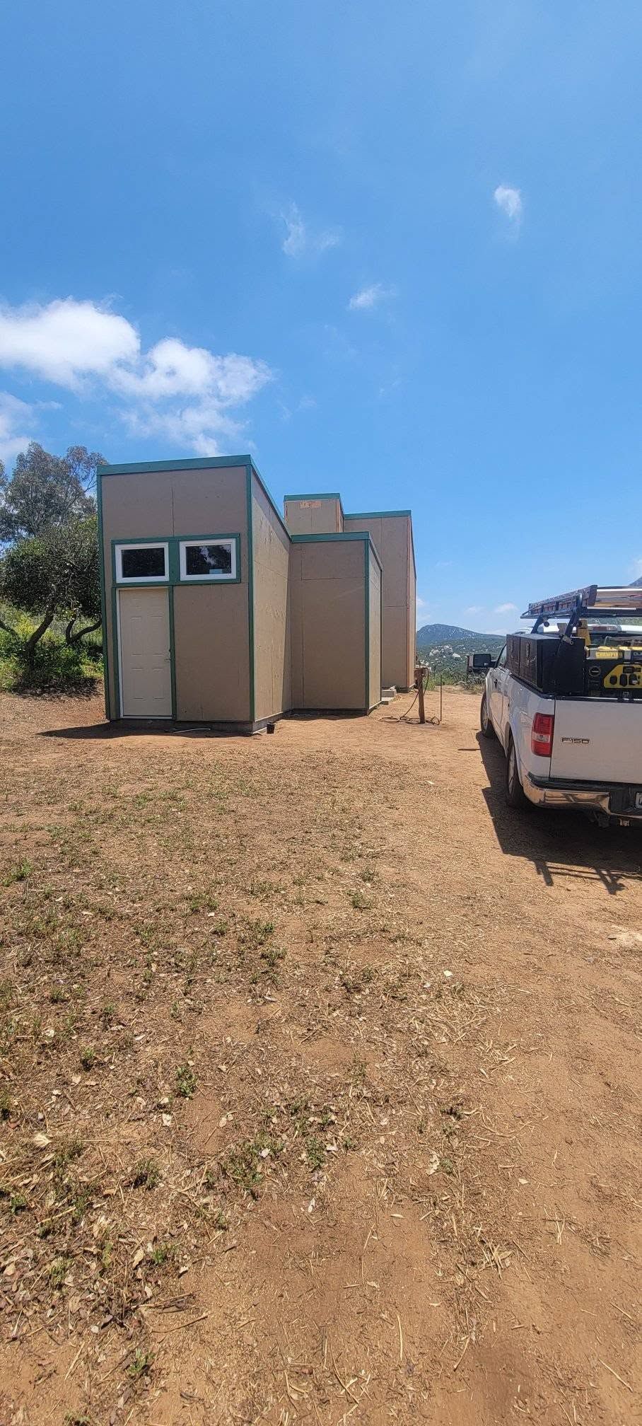 Small beige building on a dirt slope beside a white pickup truck under a bright blue sky.
