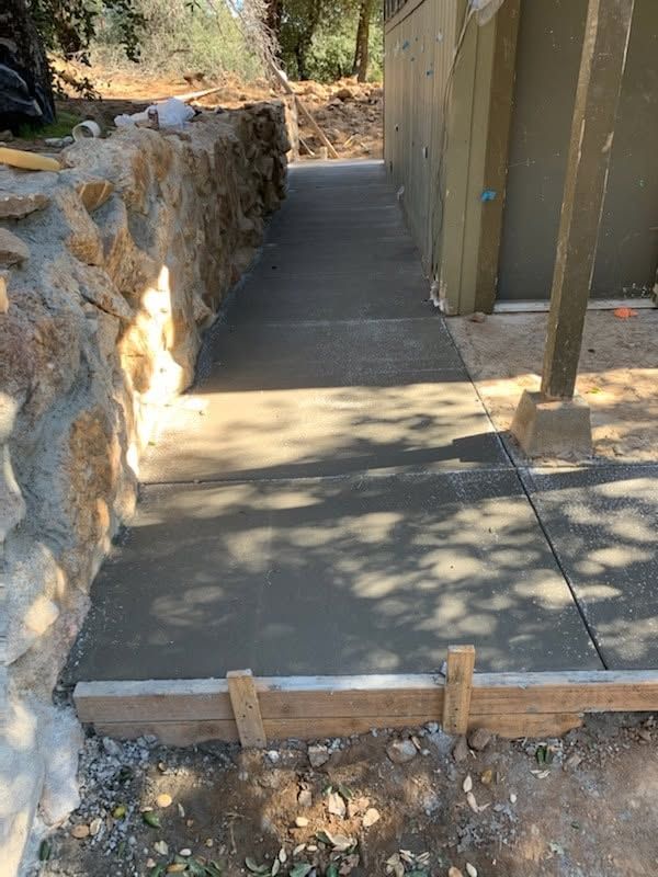 Concrete walkway with a wooden barrier at the edge beside a stone wall and fence in a sunny outdoor area