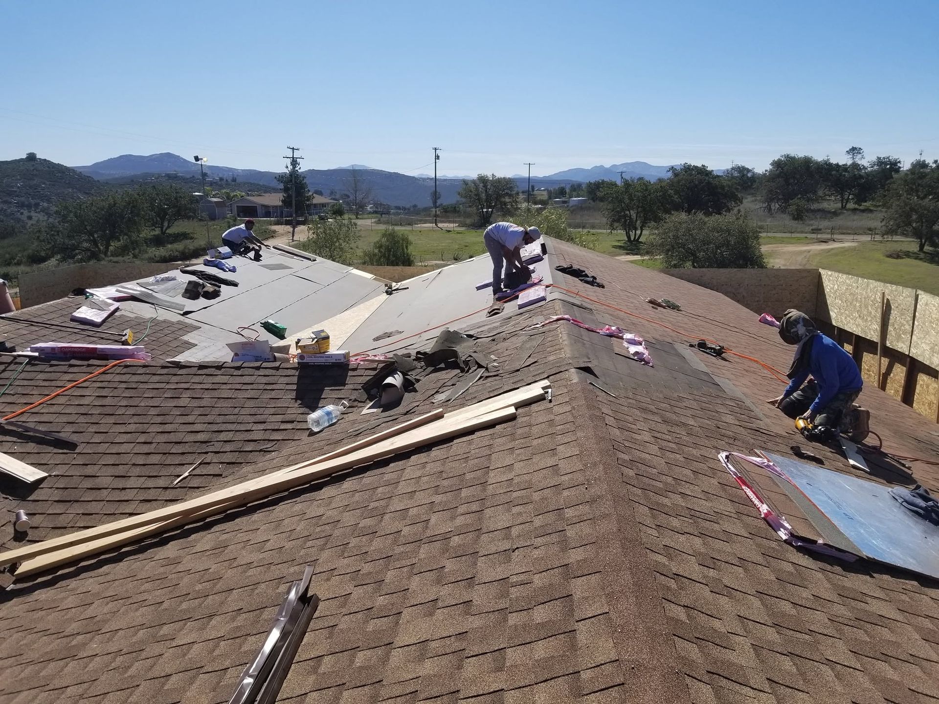 Workers repairing a shingled roof on a house, with tools and materials spread across the rooftop.