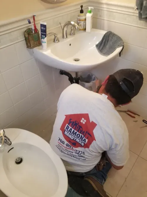 Person kneels under a bathroom sink, repairing plumbing beneath a white pedestal basin.