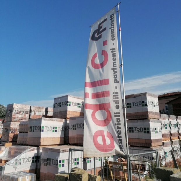 Un alto striscione di Edili.com sovrasta materiali edili accatastati sotto un cielo azzurro.