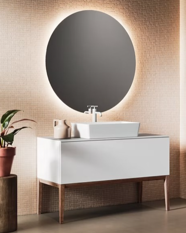 Bagno moderno con mobile lavabo bianco, specchio rotondo e parete rivestita con piastrelle a mosaico color beige tenue.