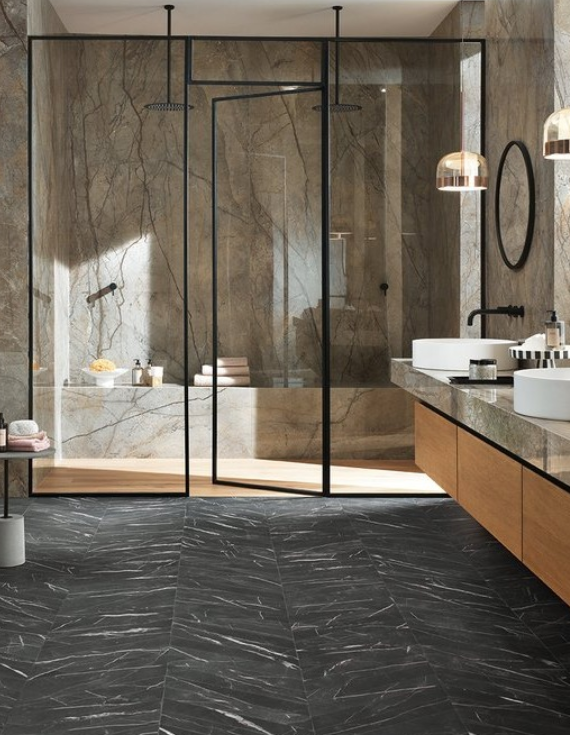 Bagno moderno con pareti in marmo, porte doccia in vetro nero e mobiletto con lavabo su pavimento piastrellato scuro.