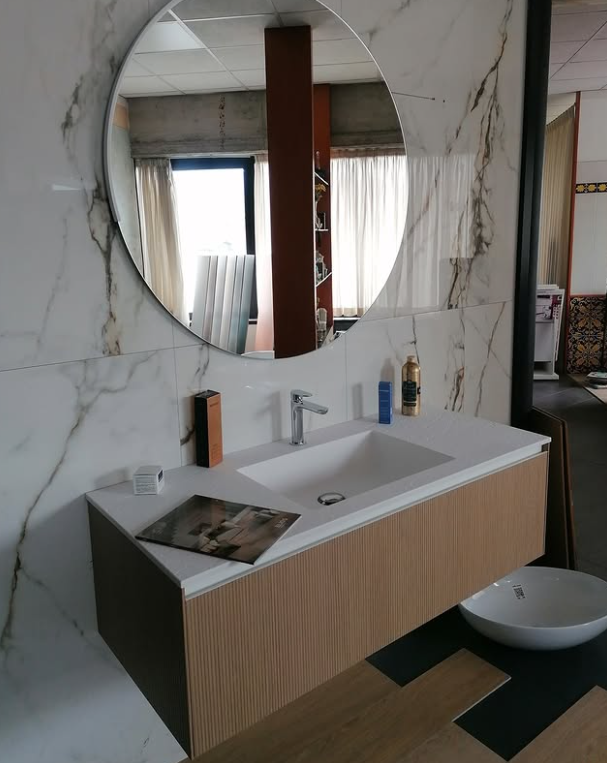 Mobile bagno moderno con specchio rotondo, lavabo bianco, parete in marmo e mobiletto con dettagli in legno.