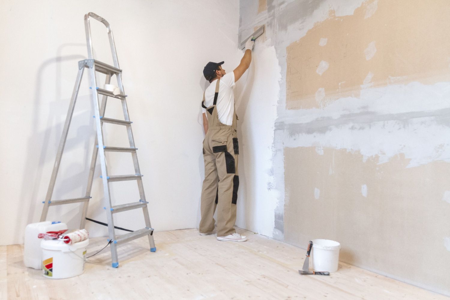 Man in work clothes on a ladder, applying plaster to a wall. Interior room with materials.