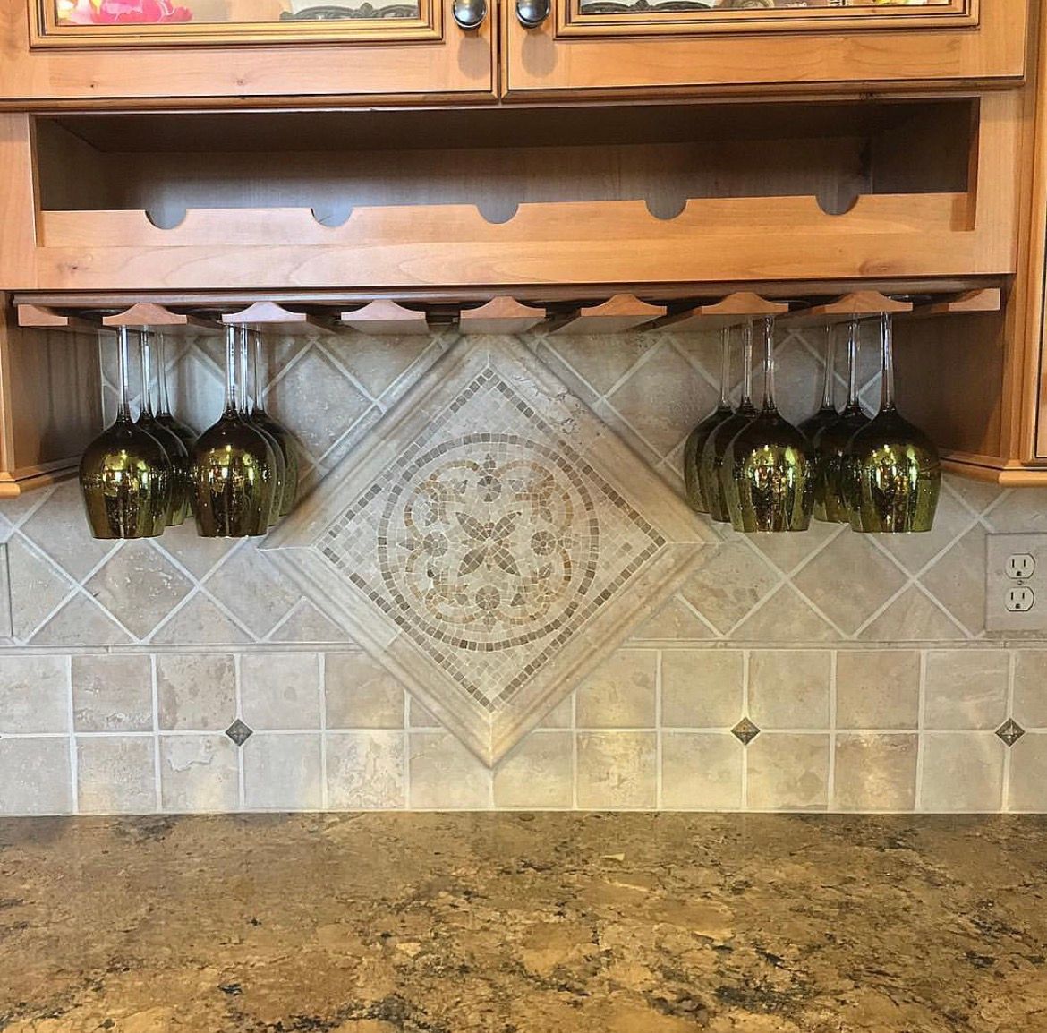 Kitchen wine glass rack with hanging green wine glasses; decorative tile backsplash, wooden cabinets.