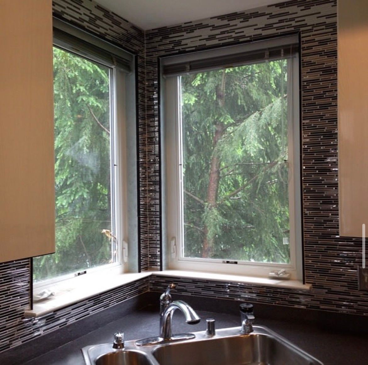 Corner kitchen windows with silver faucet and sink, black countertop, and mosaic tile. Lush green trees outside.