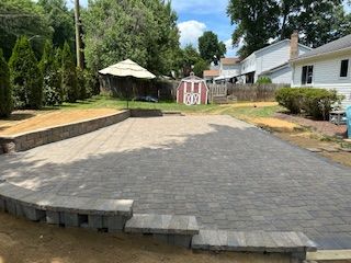 Backyard patio with stone pavers and retaining wall; shed and houses in background.