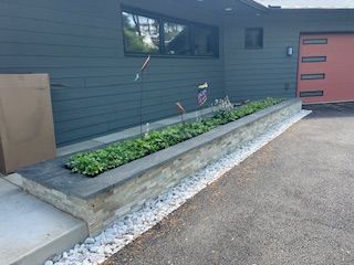Raised stone planter box with green plants next to a dark gray house with a red garage door.