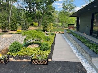 A paved walkway with garden beds of greenery and a tree alongside a dark-colored building with glass windows.