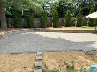 A paved patio with a retaining wall, evergreen trees, and stepping stones leading to it.