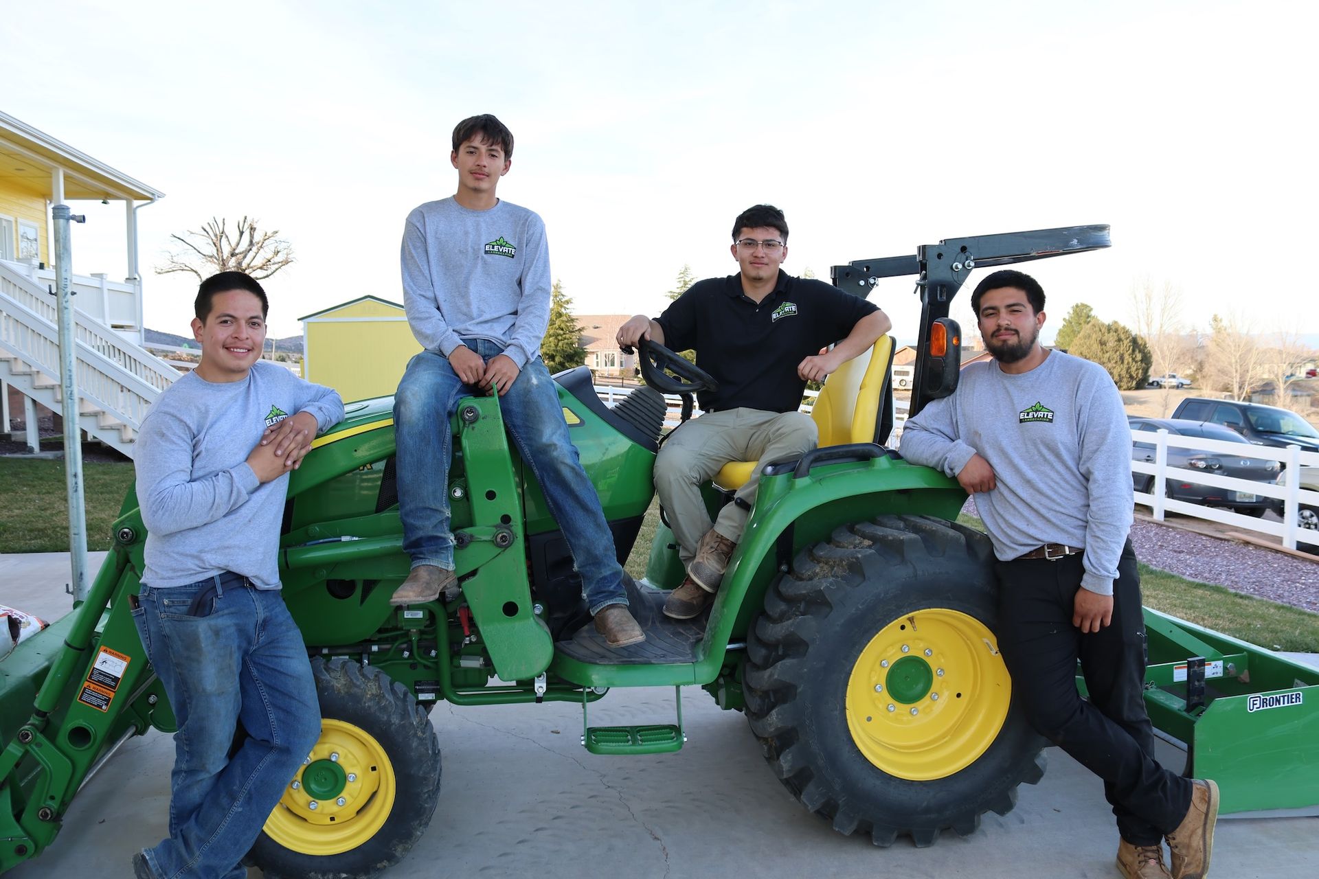 Four people pose on a green tractor; one sits at the wheel.  All wear matching shirts.  Outside, sunny.