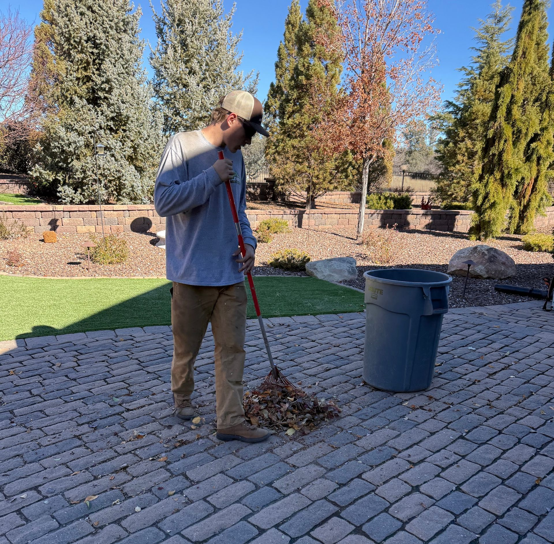 Man rakes leaves on a brick patio into a gray trash can on a sunny day.