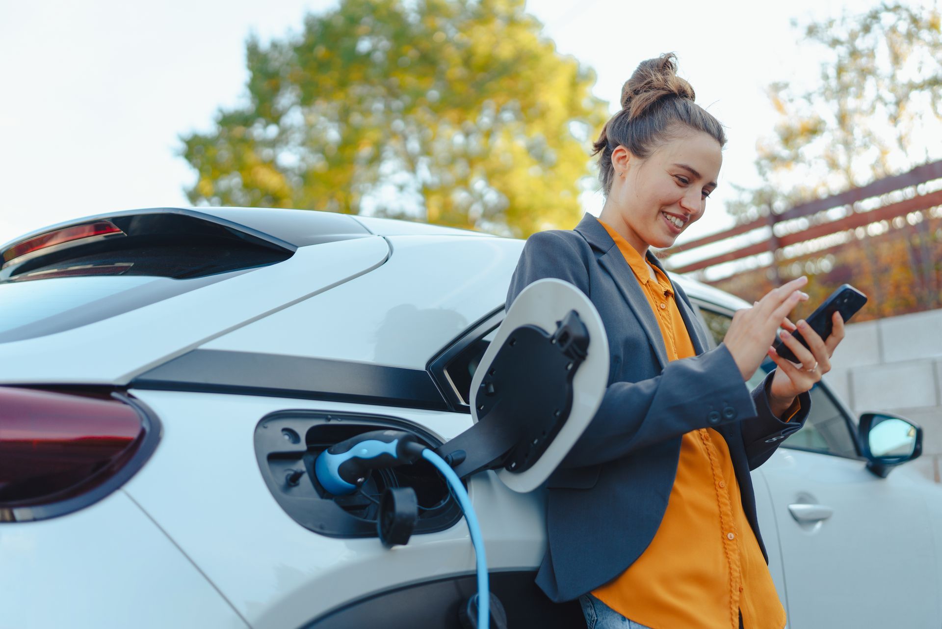 Une femme souriante vérifie son téléphone pendant que sa voiture électrique se recharge à l'extérieur.
