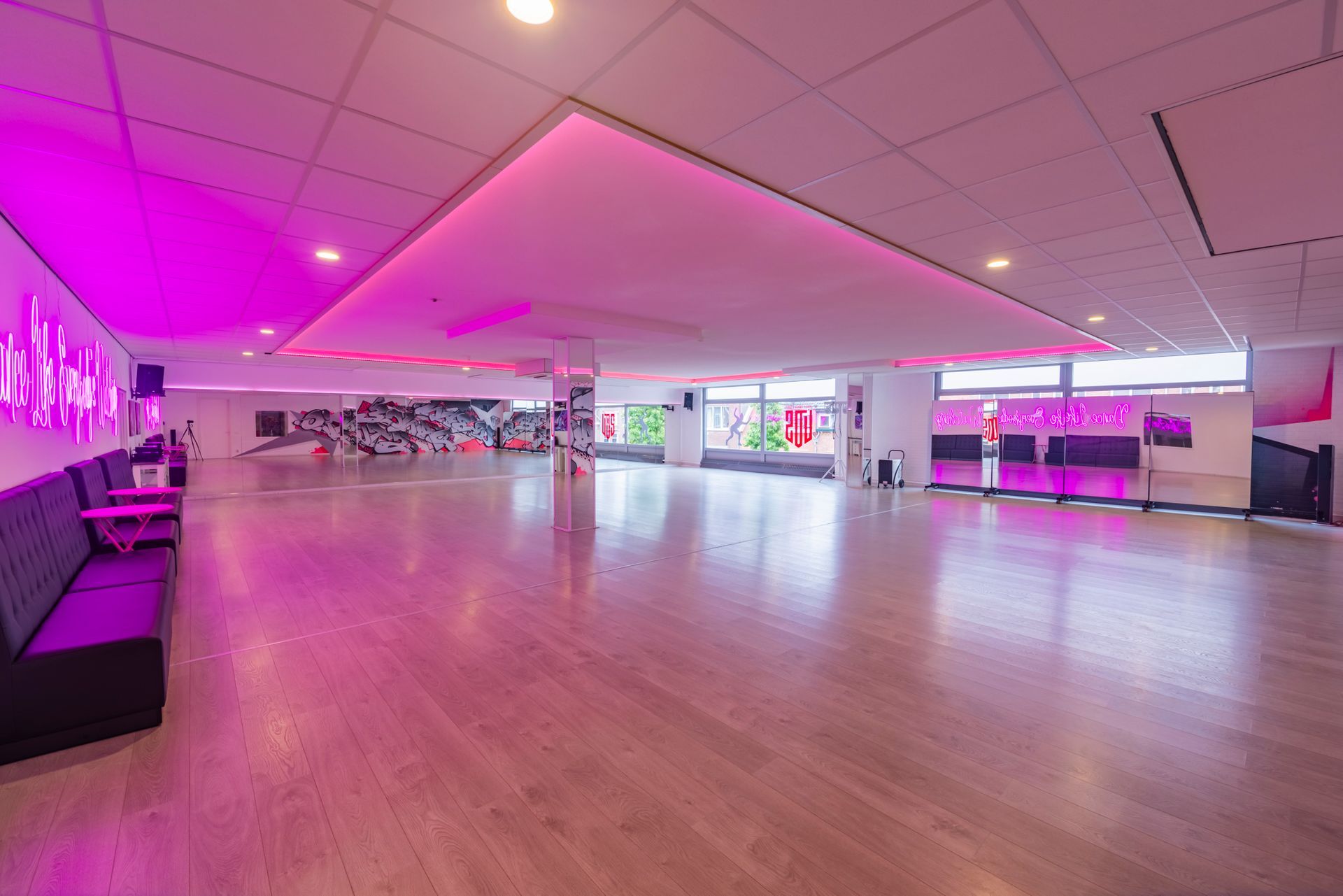 Leegstaande moderne dansstudio met roze verlichting, houten vloer, spiegels en grote ramen.